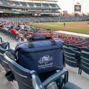 A Taiga Navy soft side 18 pack cooler from Taiga Coolers rests on a stadium seat, overlooking a baseball field as fans fill the stands. The cooler shows the Taiga Coolers logo and website.