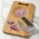 A mezzaluna knife and sliced red onions rest on the NULU Signature Cutting Board by NULU Inc., which features an integrated mincing bowl in its recessed center, all placed on a marble surface.