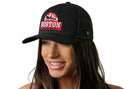 A woman with long brown hair smiles while wearing the Haka Hat Boston City On A Hill Trucker Hat, a black cityscape snapback by Haka Hat with 3D embroidery and a red and white city skyline on the front. White background.