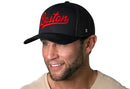 A man with a short beard and mustache smiles against a white background, wearing a beige T-shirt and the Haka Hat Boston Red Script Trucker Hat, a black Massachusetts snapback from Haka Hat.
