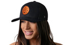 A woman with long brown hair smiles while wearing the Haka Hat Boston Trucker Hat Leather | Black BOS X Snapback, featuring a round orange leather patch with BOS and a bat and ball design. The background is plain white.