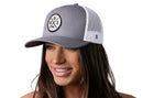 A woman with long brown hair smiles while wearing a gray and white Haka Hat Boston Trucker Hat featuring the BOS X snapback design. The background is white.