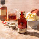 A jar of Honey Truck Co Raw Bourbon Infused Wildflower Honey sits on a table next to a bowl of vanilla ice cream, an ice cream scoop, and a St. Augustine Distillery Bourbon bottle in the background. Warm lighting highlights the dessert scene, perfectly showcasing the rich wildflower honey from Honey Truck Co infused with smooth bourbon notes.