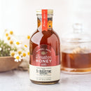 A jar of Honey Truck Co Raw Bourbon Infused Wildflower Honey sits on a white surface, with a honey dipper, glass jar, and small flowers in the background. The label highlights raw and bourbon-infused qualities, capturing the essence of rich wildflower honey with subtle bourbon notes.