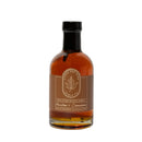 A clear glass bottle of Under The Bark Syrup Co Bourbon + Cinnamon Maple Syrup features a brown label with a maple leaf logo and the text "100% Pure Maple Syrup, Bourbon & Cinnamon.