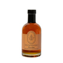 A clear glass bottle of Under The Bark Syrup Co Bourbon Maple Syrup features amber syrup, a black cap, and an orange and beige label with a maple leaf logo.