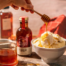 A hand drizzles rich Honey Truck Co Raw Bourbon Infused Wildflower Honey from a dipper onto vanilla ice cream in a bowl. A jar of Honey Truck Co honey, a glass of honey, and bottles of St. Augustine Distillery Bourbon are on the table, illuminated by sunlight.