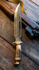 A Texan Knives Damascus Steel Vintage Bowie Knife – 13" with Brass Guard & Stag Horn Handle, with its leather sheath, rests on a wooden surface beside a rifle with a leather strap.