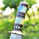 Close-up of the Texan Knives Damascus Steel Antique Bowie Knife—13", featuring a brass guard, colorful bone & pakkawood handle, and intricate patterns, set against a blurred green outdoor background. Includes leather sheath.