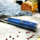 The Spaceman Knives Interstellar Bread Knife by Spaceman Knives, featuring a textured blue handle and AUS-10 steel blade, rests on stone with peppercorns, a copper-lidded jar, and blue fabric in the background.