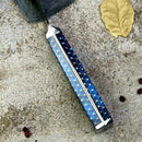 A close-up of the Spaceman Knives Interstellar Bread Knife handle, showcasing its blue, scale-like pattern on a sandy surface with a dried yellow leaf and small red berries nearby.