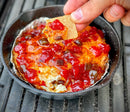 A hand holds a cracker topped with creamy cheese and Terrapin Ridge Farms Hot Pepper Bacon Jam, above a skillet of baked cheese smothered in chunky red jam—Terrapin Ridge Farms’ perfect addition to charcuterie boards or snack time.