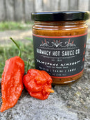 A jar of Harmacy Hot Sauce Co. Brimstone Inferno Extra Hot Tahini & Roasted Red Pepper Sauce 9oz sits on concrete beside two fresh red chili peppers, showcasing its bold Mediterranean flavors of tahini and roasted red pepper.