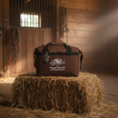 The Taiga Brown soft side 18 pack cooler by Taiga Coolers sits on a hay bale in a sunlit wooden barn, featuring a fishing logo, slogan, and website printed on the front.