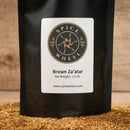 A black Spice Wheel Brown Za'atar 1/2 lb bag stands on a wooden surface, with loose brown za’atar spice blend rich in sumac scattered at its base. The bag features the Spice Wheel brand name and product label.