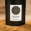 A black pouch of Spice Wheel Brown Za’atar 1 lb from Spice Wheel stands on a wooden surface, its label clearly visible amid scattered brown za’atar blend at the base. Flecks of sumac add vibrant contrast to the aromatic mix, showcasing the rich character of this traditional seasoning.