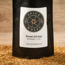 A black Spice Wheel bag labeled "Spice Wheel Brown Za’atar 1/4 lb" from the brand Spice Wheel and featuring www.spicewheel.com stands on a surface covered in aromatic brown za’atar—a signature Middle Eastern spice blend known for its earthy herbs and tangy sumac.