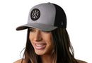 A smiling woman wears the Haka Hat Buffalo Trucker Hat in gray-black, featuring a round BUF X patch with crossed lines and the letters U, B, F, and A. The background is plain white.