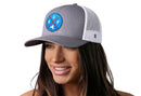 A woman with long brown hair smiles while wearing the Haka Hat Buffalo Trucker Hat | Blue Red BUF X, a gray-white snapback by Haka Hat. The hat, featuring a blue, red, and white circular patch, stands out against a plain white background.