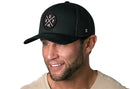A man with short brown hair and a trimmed beard smiles in a beige t-shirt and a Haka Hat Buffalo Trucker Hat | Black BUF X Snapback by Haka Hat, featuring a round embroidered patch. The background is plain white.