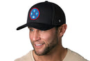 A man with a trimmed beard and light shirt smiles while wearing a black Haka Hat Buffalo Trucker Hat | Black BUF X Football Snapback, which features a blue and red circular Buffalo-themed logo. The background is white.