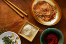 Four bowls filled with various spices and herbs, including World Spice Bulgogi Spice by World Spice, sesame seeds, and vibrant gochugaru, sit on a wooden surface with a pair of chopsticks nearby.