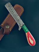 The Texan Knives Bull Cutter Handmade Damascus Steel Knife features a vibrant red, green, and beige pakkawood handle and comes with a brown leather sheath accented with yellow stitching and a button snap.