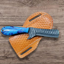 The Texan Knives Bull Cutter Knife - Custom Handmade Precision Tool features a textured J2 steel blade and blue patterned handle, resting on a tan leather sheath atop a wood-grain surface.