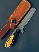 The Bull Cutter Knife by Texan Knives features a handmade Damascus steel blade and pakkawood handle, resting beside a brown leather sheath with yellow stitching, set against a dark background.