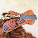 The Texan Knives Bull Knife features an 8-inch custom handmade J2 steel blade and a black, white, and yellow marbled resin handle, resting on a brown leather sheath with blue accents and intricate designs atop a fur surface.