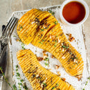 Sliced, roasted butternut squash garnished with herbs, walnuts, and SALTOPIA MAPLE ME CRAZY Maple Garlic & Thyme Salt on a white tray, served with cutlery and a bowl of sauce.