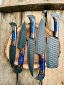 Five knives from the Texan Knives Handmade Damascus Steel Chef Knife Set, with textured black blades and brown wood grain handles accented by blue resin, are arranged on a round wooden surface against a rustic wooden background.