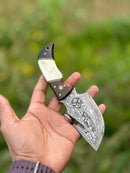 A hand holds the Texan Knives Custom Handmade Damascus Steel Guthook Skinning Knife, featuring a 7.5” blade and a rosewood, camel bone & black sheet handle, with leather sheath—shown against a blurred green outdoor background.