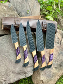Five Texan Knives chef knives from the Best Knife Block Set, featuring 13" forged stainless steel blades and olivewood & epoxy resin handles with leather sheaths, are displayed on rocks with greenery and a brown leather case in the background.