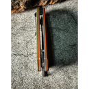 Close-up top view of the Spaceman Knives Galatea Folding Pocket Knife by Spaceman Knives, partially open to reveal its blade spine, textured grip, and orange and green handle accents on a gray stone-textured surface.