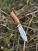 The Texan Knives Hunting Blade – a 10" Damascus steel knife with a rosewood handle, brass guard, and leather sheath – rests outdoors atop dried twigs and leaves.