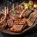 A wooden platter holds grilled sausages, ribs, and lamb chops, garnished with rosemary and SALTOPIA Infused Sea Salts’ Saltopia Ultimate Carnivore Salts, served beside grilled potatoes, zucchini, and onions on a dark surface.