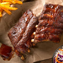 Barbecue pork ribs glazed in sauce rest on brown parchment, served with fries dusted with SALTOPIA Infused Sea Salts—the Ultimate Carnivore Salts—and a side of coleslaw. A basting brush lies nearby.
