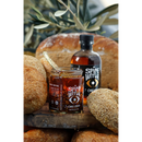 A jar of Spice Witch Chili Oil + Chili Crisp with a spoon inside is surrounded by bread, while a bottle of the same product sits on a rustic wooden board, framed by leafy branches in the background.