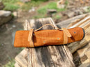 A Texan Knives Sharp Kitchen Knife Set with a 13" Damascus steel blade, olivewood handle, and leather sheath rests outdoors on weathered wood; grass and blurred logs provide rustic charm in the background.