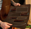 A person in an apron displays the Texan Knives Best Dishwasher Safe Knife Set—13" forged Damascus steel blades with bone & Pakkawood handles in a leather sheath. Fresh carrots and greens peek from the background, making this a kitchen essential.