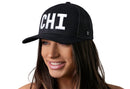 A smiling woman wears the Haka Hat Chicago White Block Trucker Hat, a black Illinois snapback with bold white CHI letters on the front; the background is plain white.
