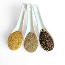 Three white ceramic spoons hold yellow, light brown, and dark brown seasonings, side by side on a white background—ideal for highlighting the SALTOPIA Kitchen Favorite Flavors Gift Set: COOKS SURVIVAL KIT infused sea salts.