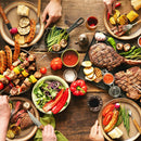A lively meal for four unfolds on a rustic wooden table, featuring grilled meats, vegetables, salad, and sauces enhanced by SALTOPIA Infused Sea Salts from the Saltopia Kitchen Favorite Flavors Gift Set: COOKS SURVIVAL KIT.