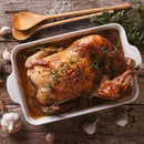 A roasted whole chicken sits in a white baking dish, surrounded by garlic and herbs, featuring SALTOPIA Infused Sea Salts from the Saltopia Kitchen Favorite Flavors Gift Set: COOKS SURVIVAL KIT on a rustic wooden table.