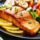 Grilled salmon fillet garnished with SALTOPIA Infused Sea Salts from the Saltopia Kitchen Favorite Flavors Gift Set: COOKS SURVIVAL KIT, served with lemon slices, fresh salad, and seasoned potatoes on a black plate.