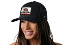 A smiling woman with long brown hair wears the Haka Hat California Flag Trucker Hat, a black CA Snapback featuring a California Republic bear patch on the front, against a plain white background.