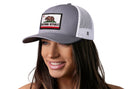 A woman smiles and looks to the side, wearing a Haka Hat California Flag Trucker Hat in gray and white with a state bear patch.