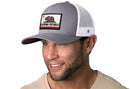 A smiling man in a beige shirt wears the Haka Hat California Flag Trucker Hat (Gray-White CA Snapback) by Haka Hat, featuring a bear and red star patch, standing against a white background.