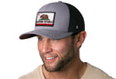 Smiling man with a short beard wears a Haka Hat California Flag Trucker Hat (Gray-Black CA Snapback) and a light beige shirt, set against a white background.